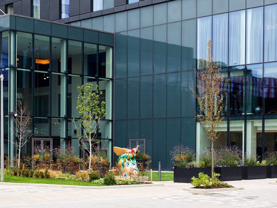 A modern building with large glass windows, a landscaped garden with various plants, and a colorful sculpture in the foreground.