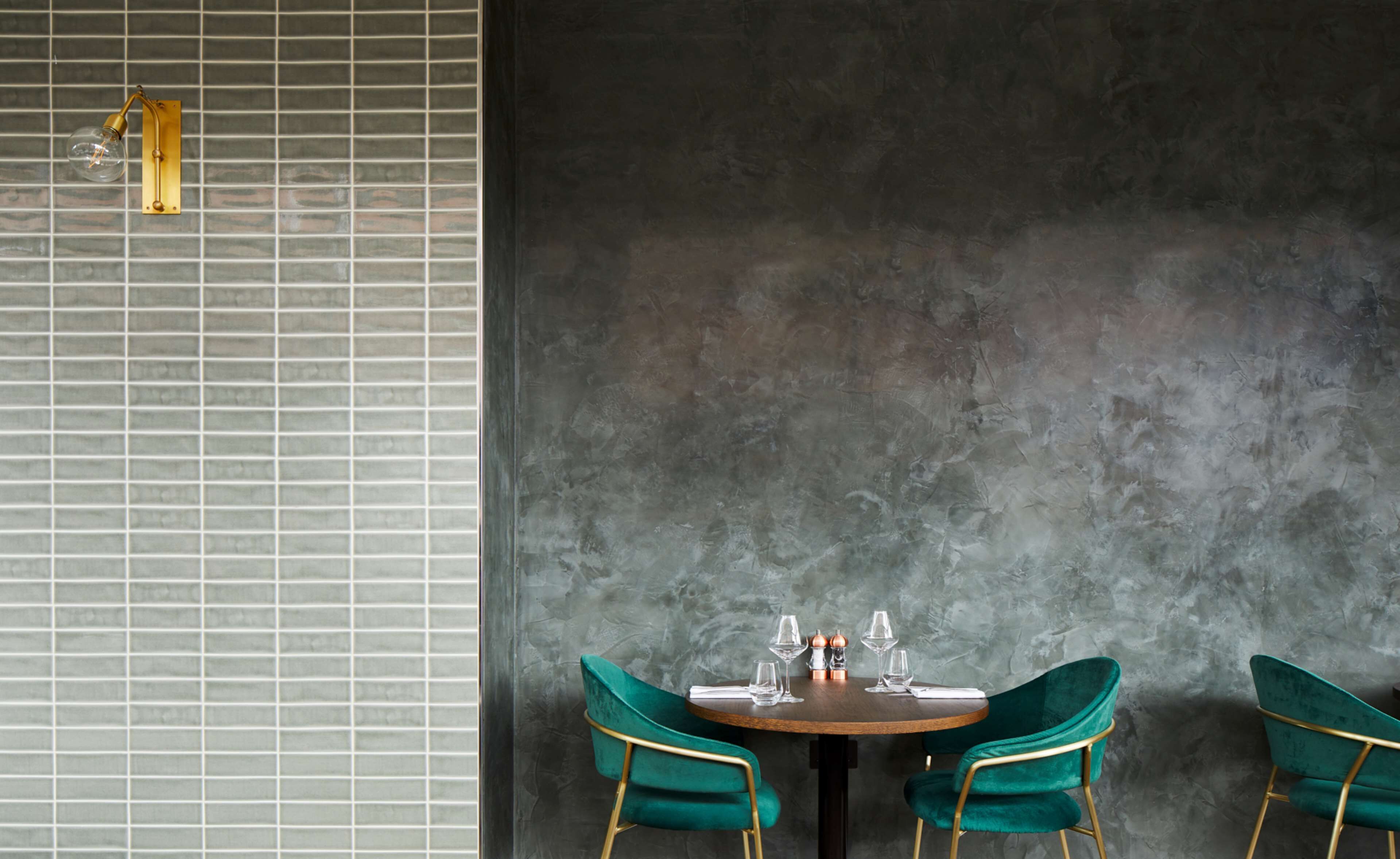 A dining area with a round table, two green velvet chairs, and a wall with a grid pattern and a light fixture.