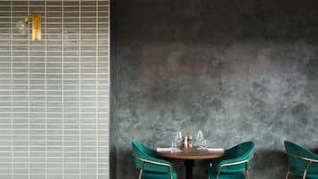 A dining area with a round table, two green velvet chairs, and a wall with a grid pattern and a light fixture.