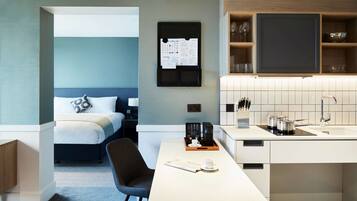 A modern hotel room with a bed, a desk with a coffee maker, a TV, and a kitchen area with a sink and cabinets.