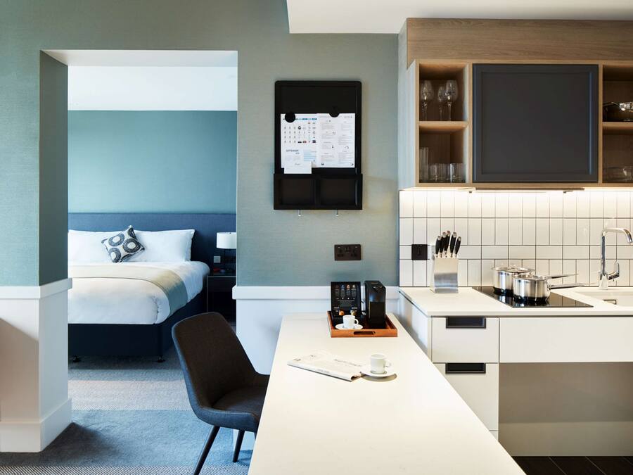 A modern hotel room with a bed, a desk with a coffee maker, a TV, and a kitchen area with a sink and cabinets.