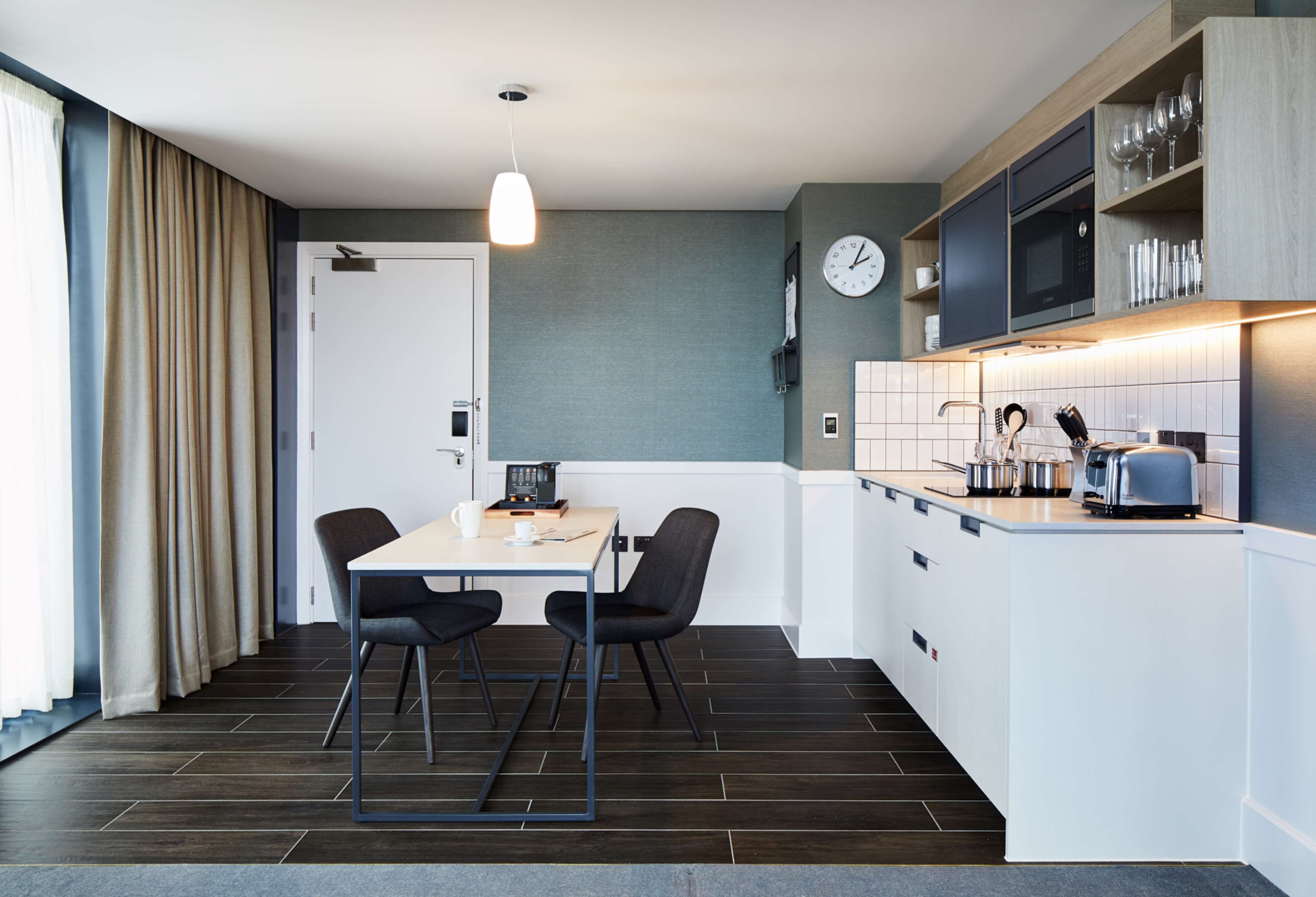Eine moderne Küche mit Essbereich, ausgestattet mit einer weißen Arbeitsplatte, dunklem Holzboden und einer Pendelleuchte.