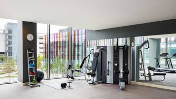 A modern gym with exercise equipment, a clock, and a view of buildings through large windows.