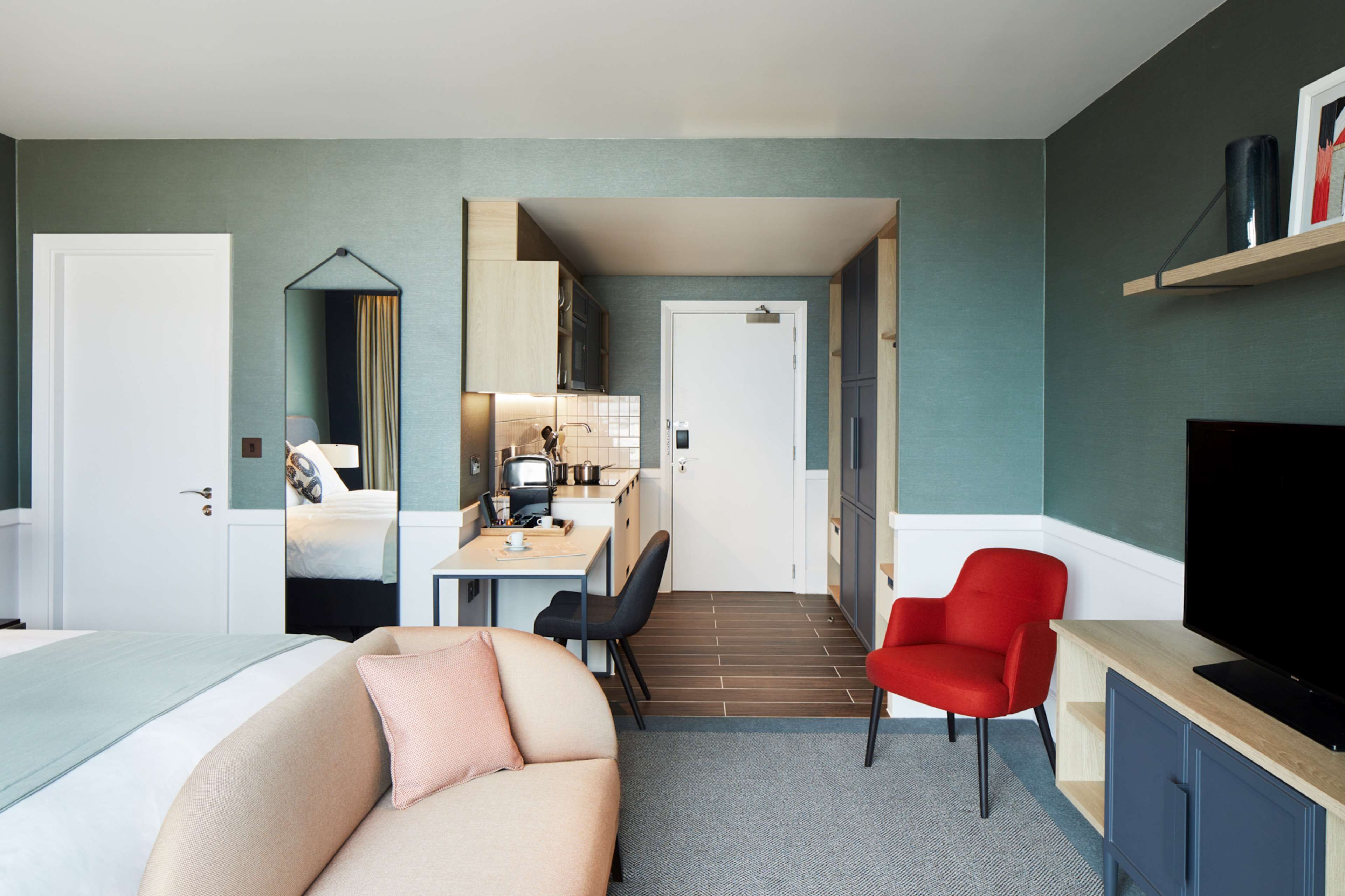 A compact hotel room with a bed, a sofa, a desk with a chair, a television, and a kitchen area.