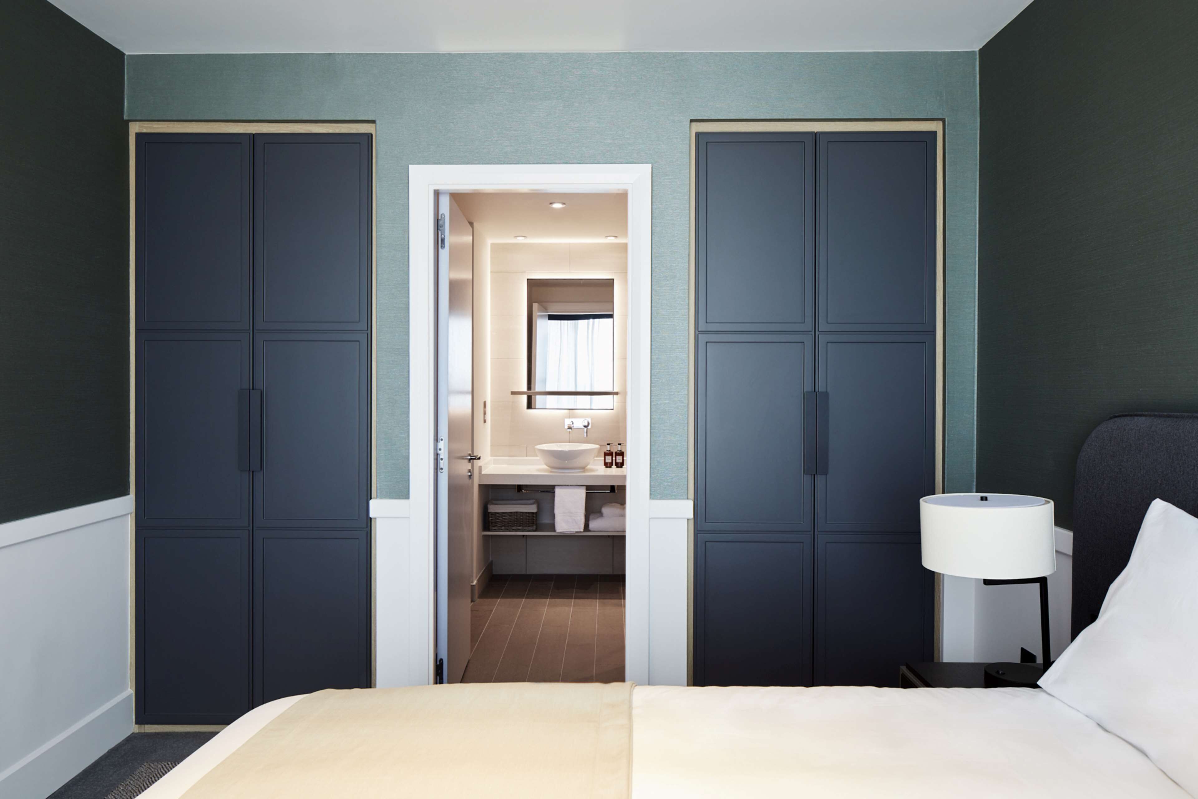 A bedroom with a bed, a nightstand, a lamp, and a view into a bathroom with a sink and mirror.