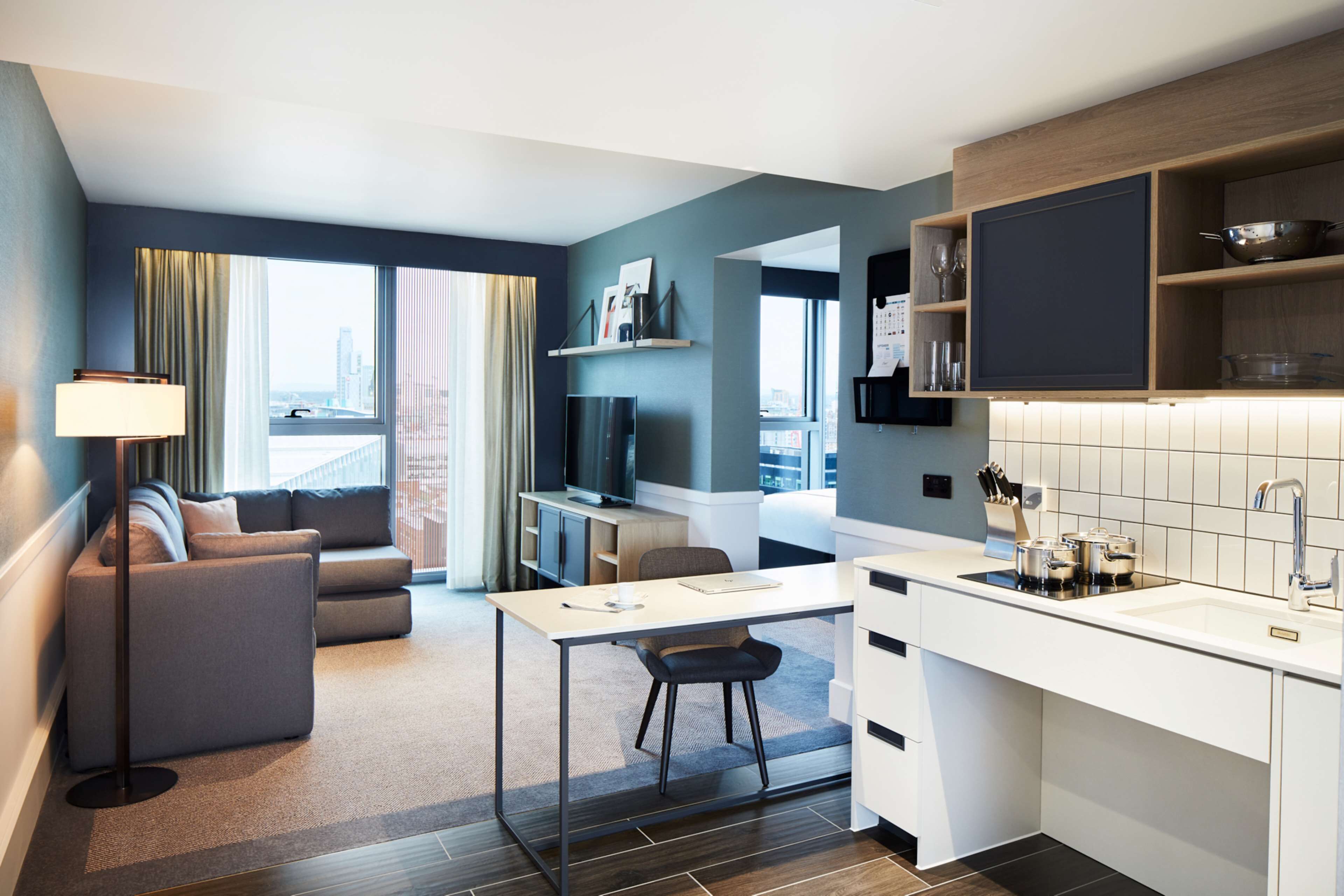 A modern hotel room with a sofa, a desk, a chair, a TV, a kitchen area with a sink and cabinets, and a large window offering a city view.