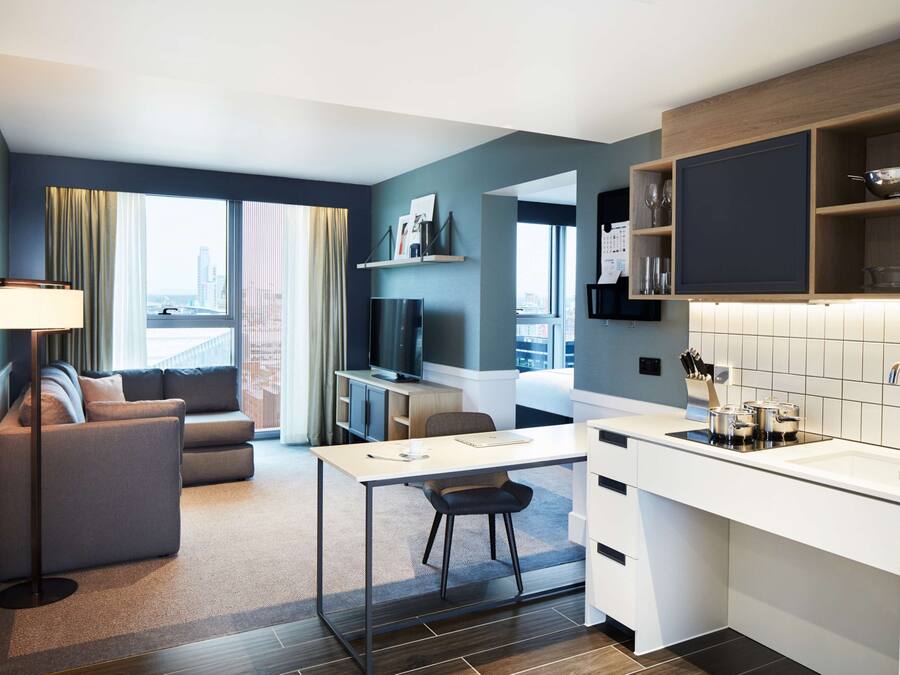A modern hotel room with a sofa, a desk, a chair, a TV, a kitchen area with a sink and cabinets, and a large window offering a city view.