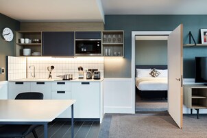 Cocina compacta con armarios blancos, microondas y cafetera. Se ve una cama con almohada a través de una puerta abierta.