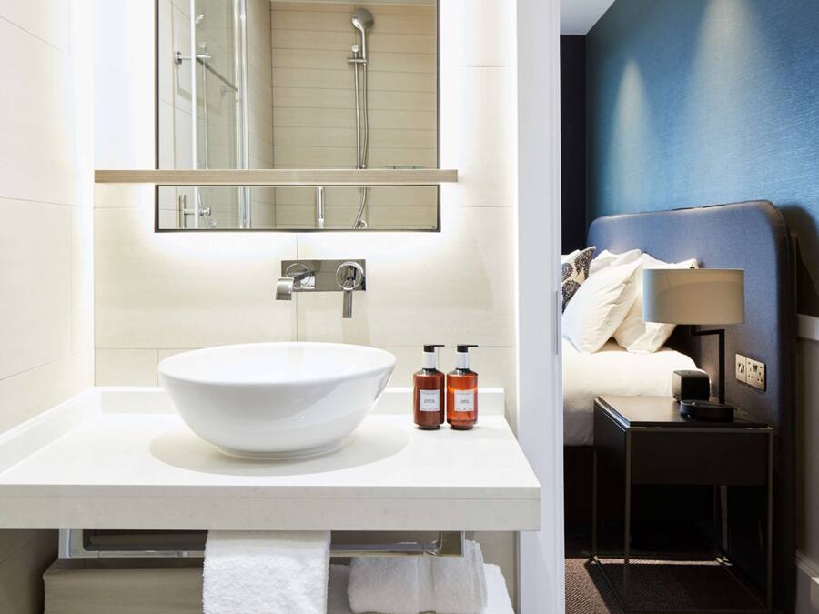 Un baño moderno con un lavamanos blanco grande, un espejo encima y una ducha visible en el reflejo. Hay una mesita de noche pequeña con una lámpara y una cama con almohadas.