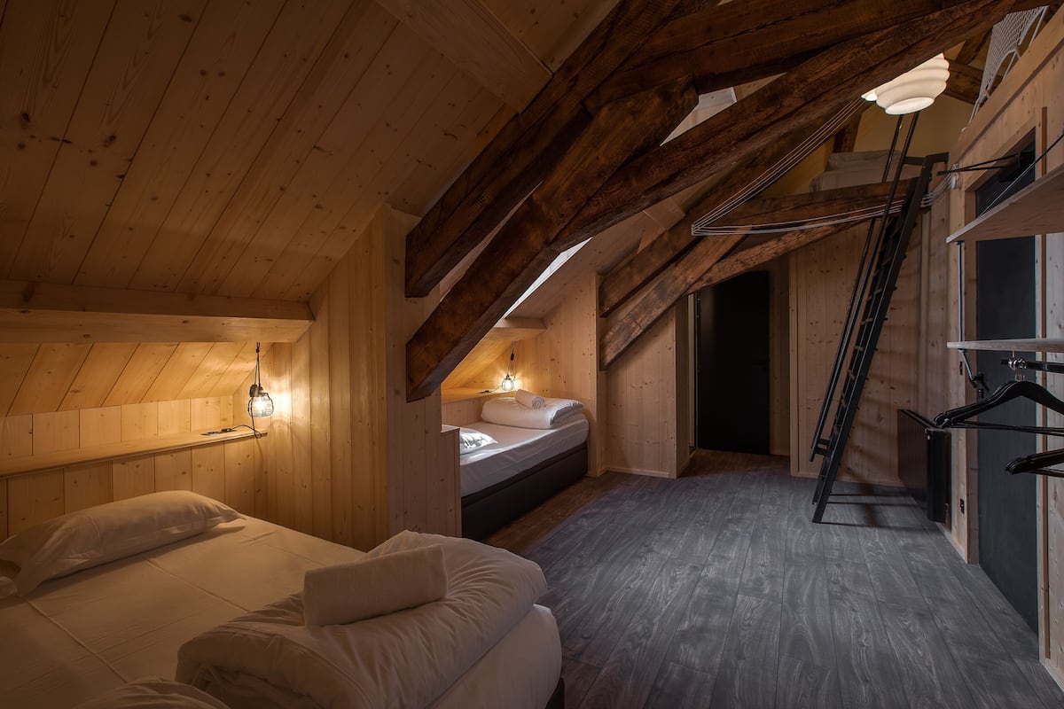 Une chambre mansardée chaleureuse avec des poutres en bois, un lit en mezzanine, un petit bureau et une échelle menant à une zone surélevée.