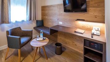 Habitación de hotel moderna con cabecera de madera, televisión de pantalla plana, una pequeña mesa redonda con una botella y vasos, un sofá y una mesita de noche con un teléfono y un control remoto.