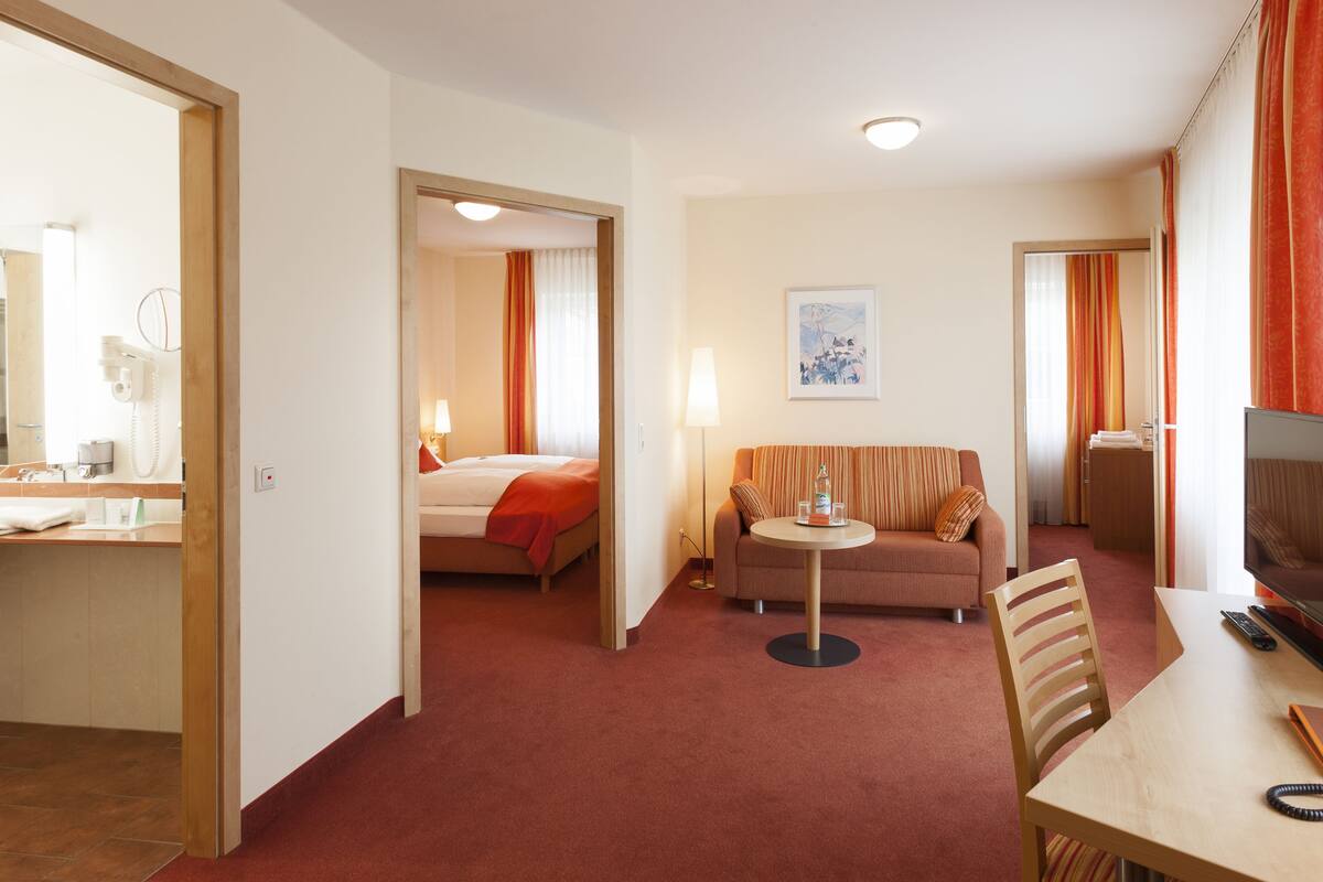 Habitación de hotel con cama, sofá, una mesita con botella y vasos, televisión y escritorio con teléfono y control remoto.