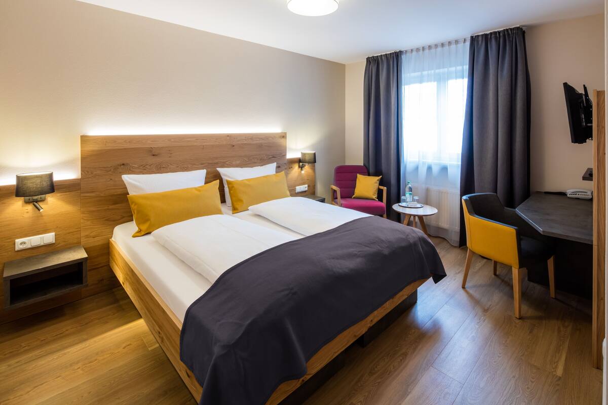 Una habitación de hotel con cama, mesitas de noche, escritorio, silla y ventana con cortinas.
