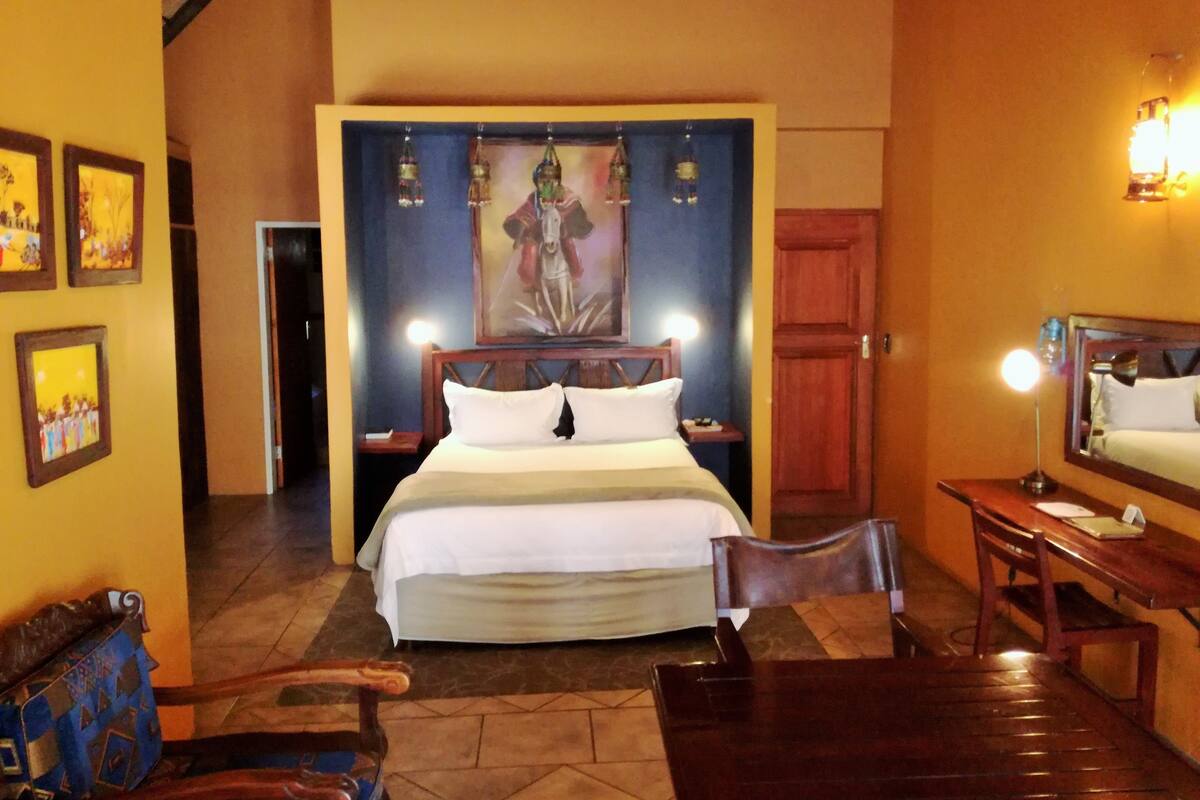 A hotel room with a bed, bedside tables, a desk, a chair, and a painting on the wall.