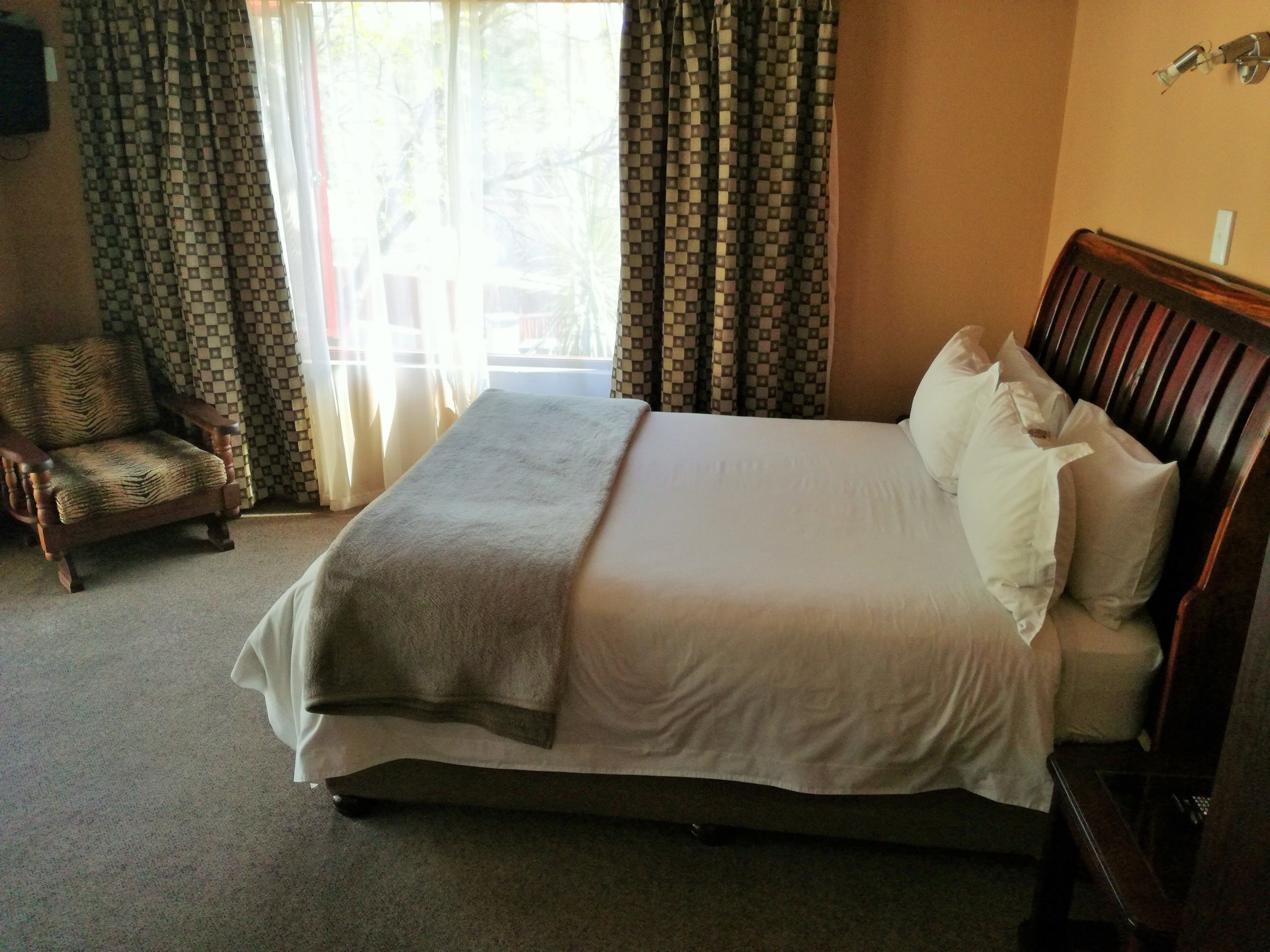 A neatly made bed with a wooden headboard, a chair with a patterned cushion, and a window with patterned curtains.