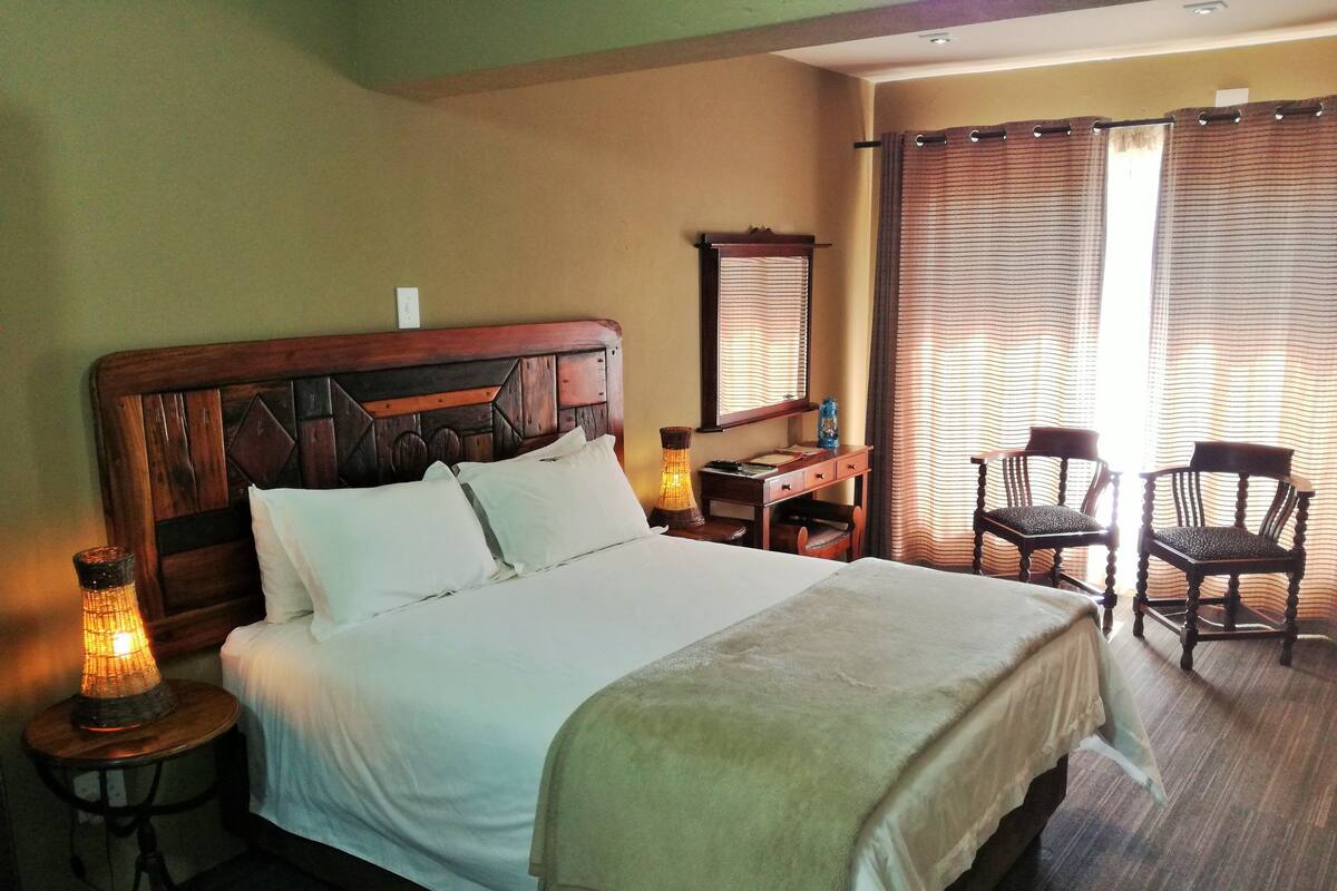 A neatly made bed with white linens, a wooden headboard, and a bedside table with a lamp. There are two wooden chairs and a desk with a mirror.