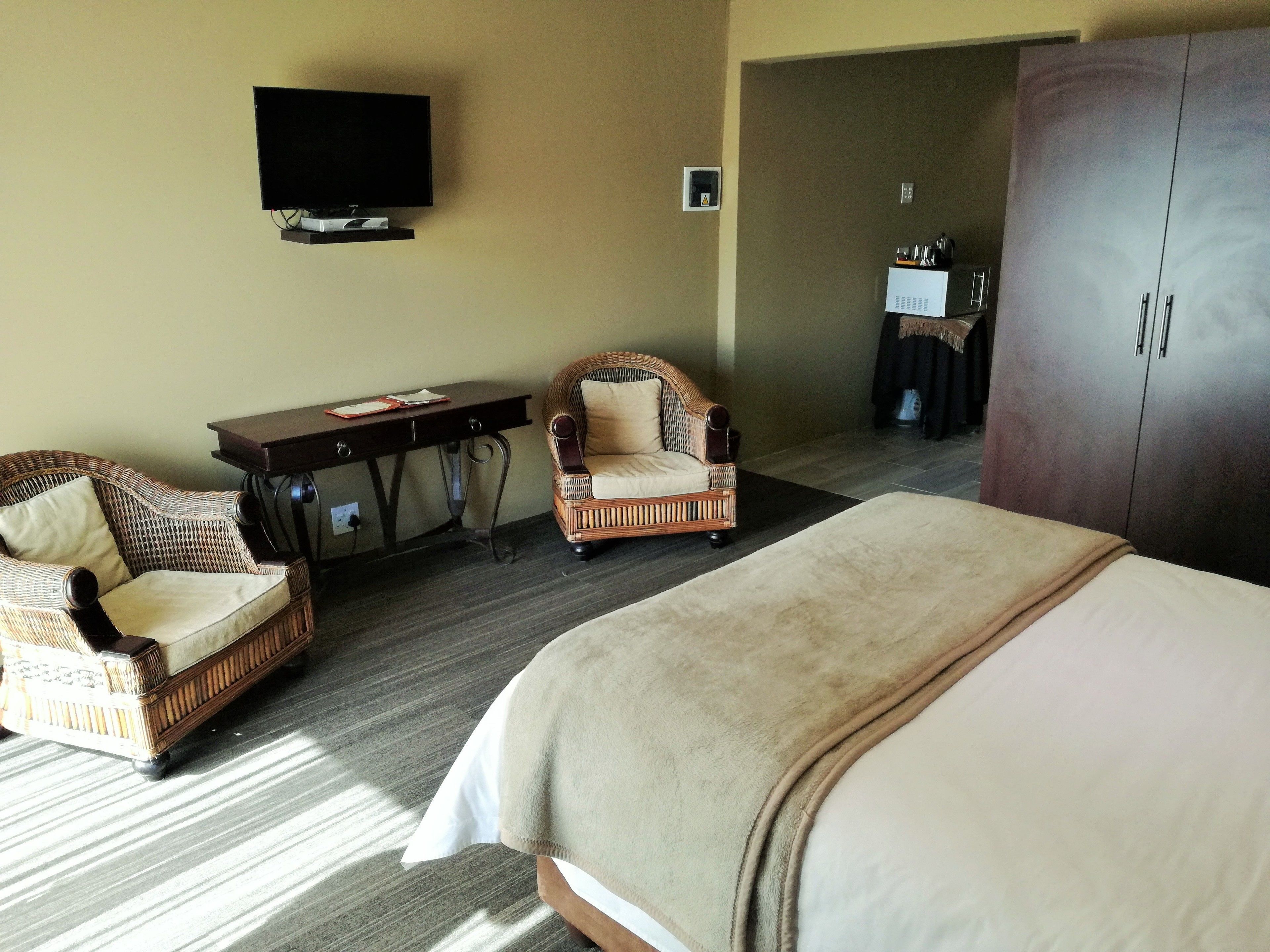 A hotel room with a bed, a wicker armchair, a desk with a flat-screen TV, and a wardrobe.