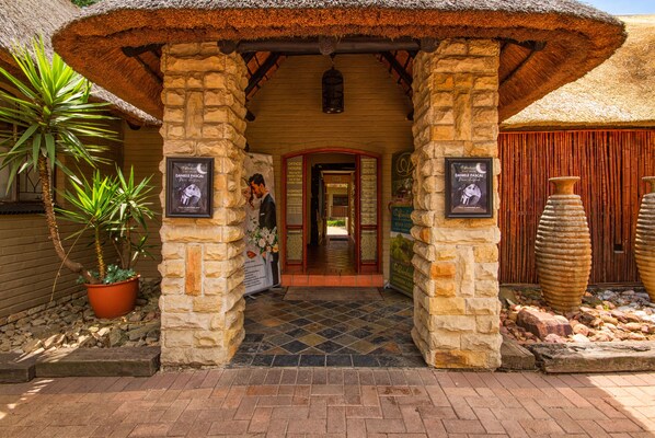 A stone building entrance with a thatched roof, potted plants, and stone pillars. - Africlassic River Lodge- Rivonia (Sandton)