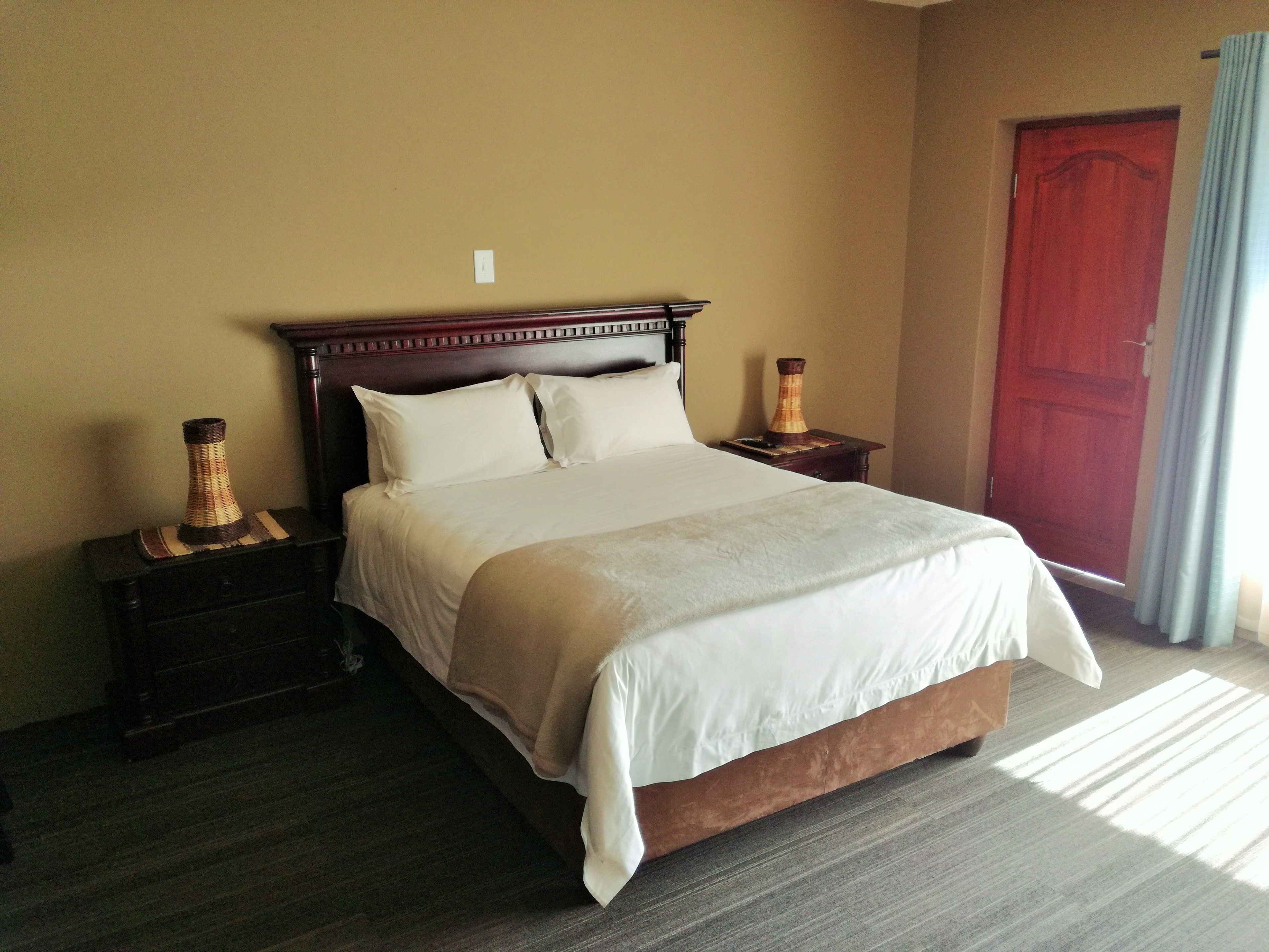 Cama bem arrumada em quarto de hotel, com cabeceira de madeira, duas mesas de cabeceira e vasos decorativos, e uma porta vermelha.