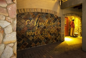 A stone wall with the text "Africlassic River Lodge Rivonia" and a statue of a person in a hat and coat. - Africlassic River Lodge- Rivonia (Sandton)