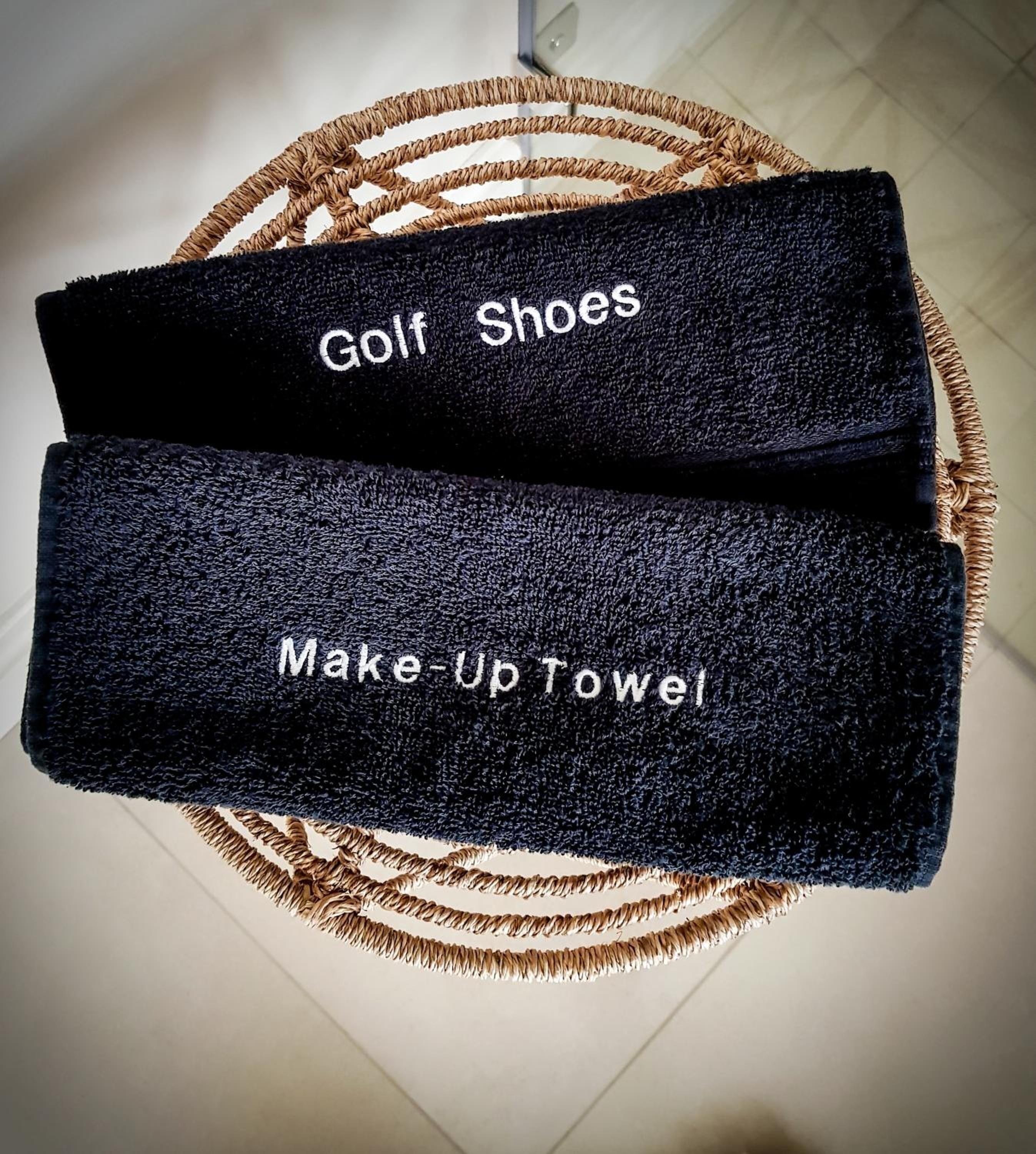 Una toalla negra con bordados que dicen "Golf Shoes" y "Make-Up Towel", colgada de una silla de mimbre.