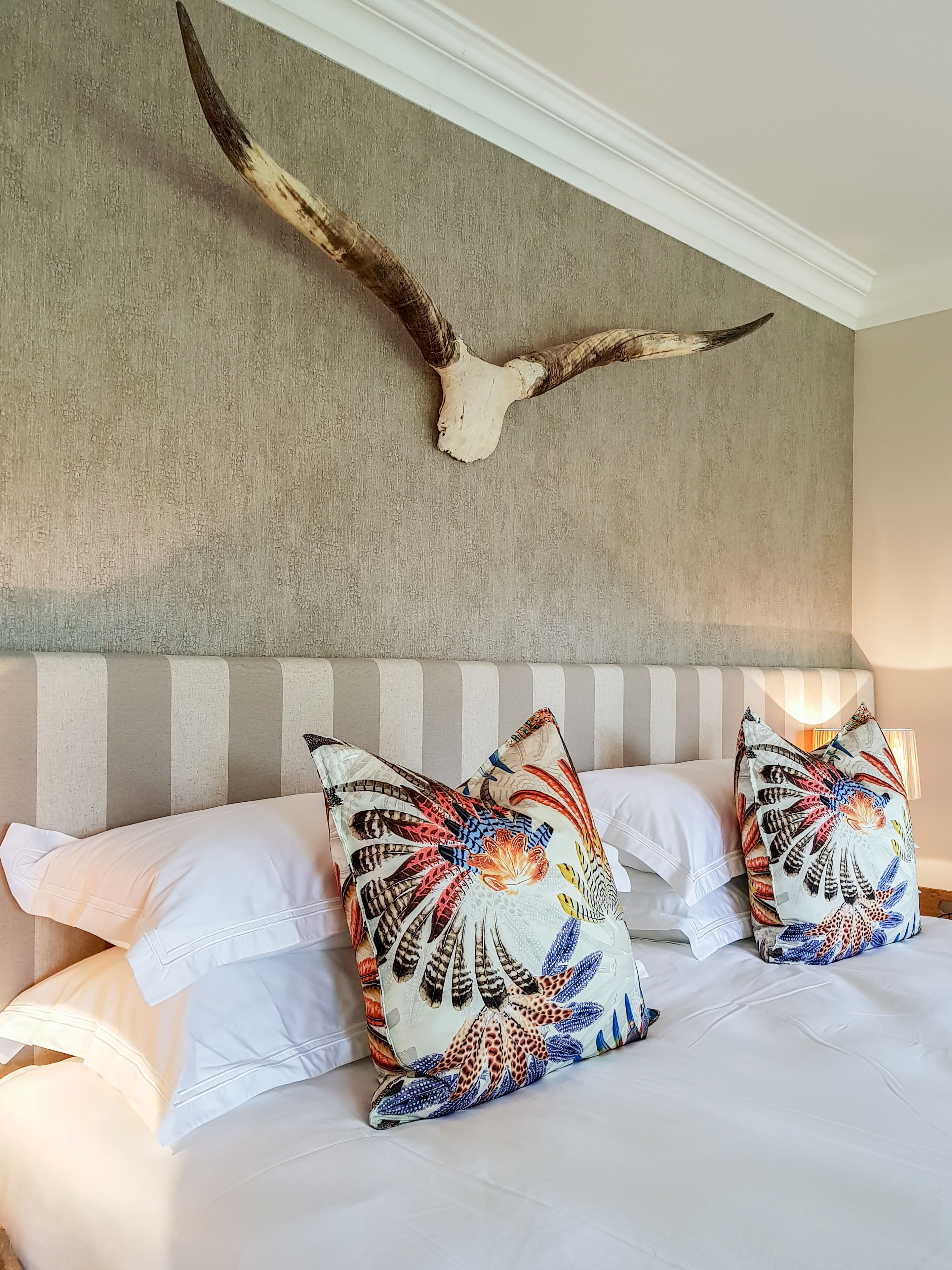 Ein Bett mit weißen Laken und farbenfrohen, gemusterten Kissen, ein Kopfteil mit vertikalen Streifen und ein großes, an der Wand befestigtes Tiergeweih.