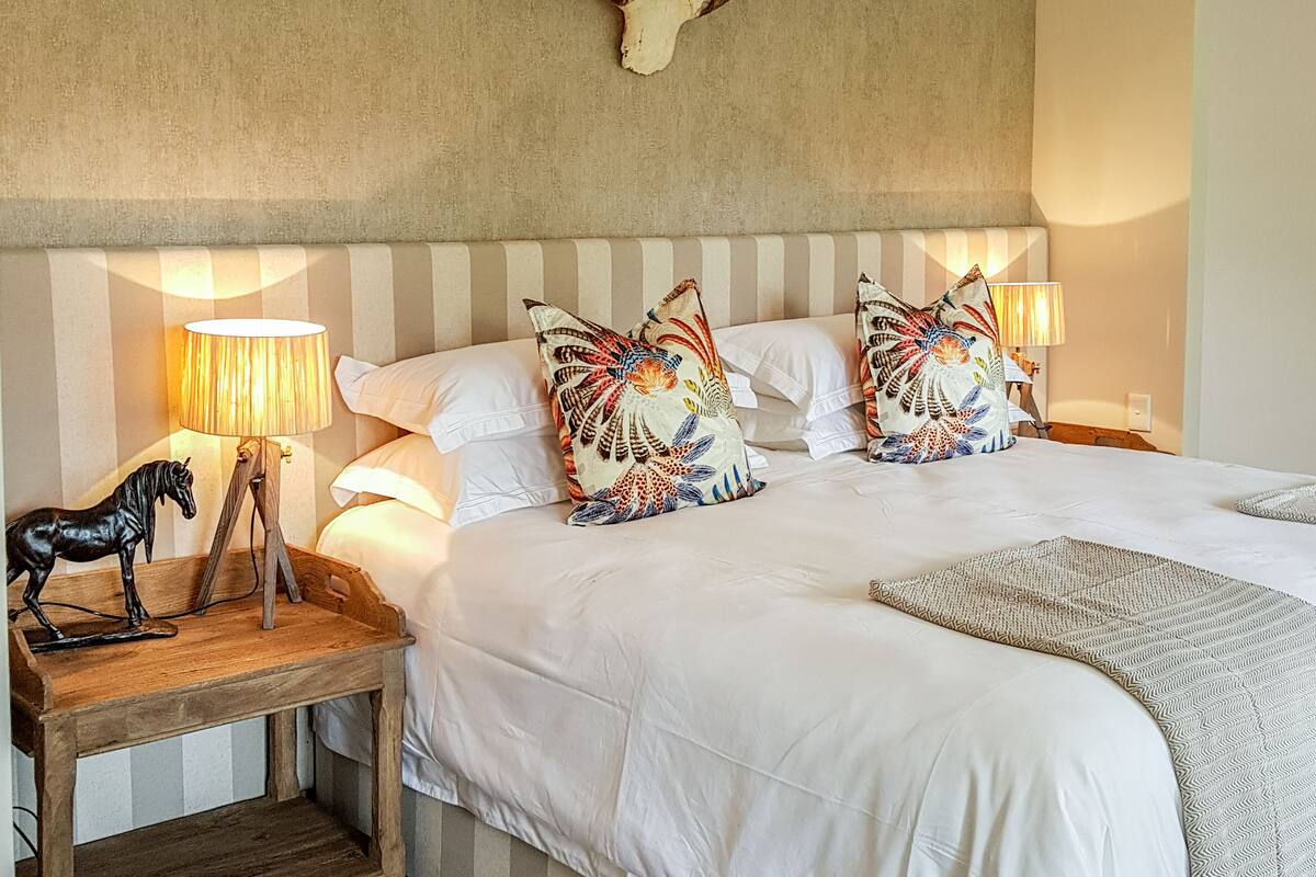 A bedroom with a large bed, striped headboard, decorative pillows, a wooden nightstand with a horse figurine, and a wall-mounted antler.