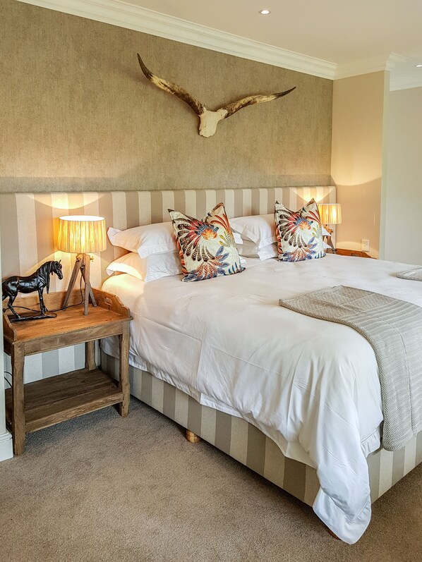 Une chambre à coucher avec un grand lit, une tête de lit à rayures, des coussins décoratifs, une table de chevet en bois ornée d’une figurine de cheval et une ramure de cerf fixée au mur.