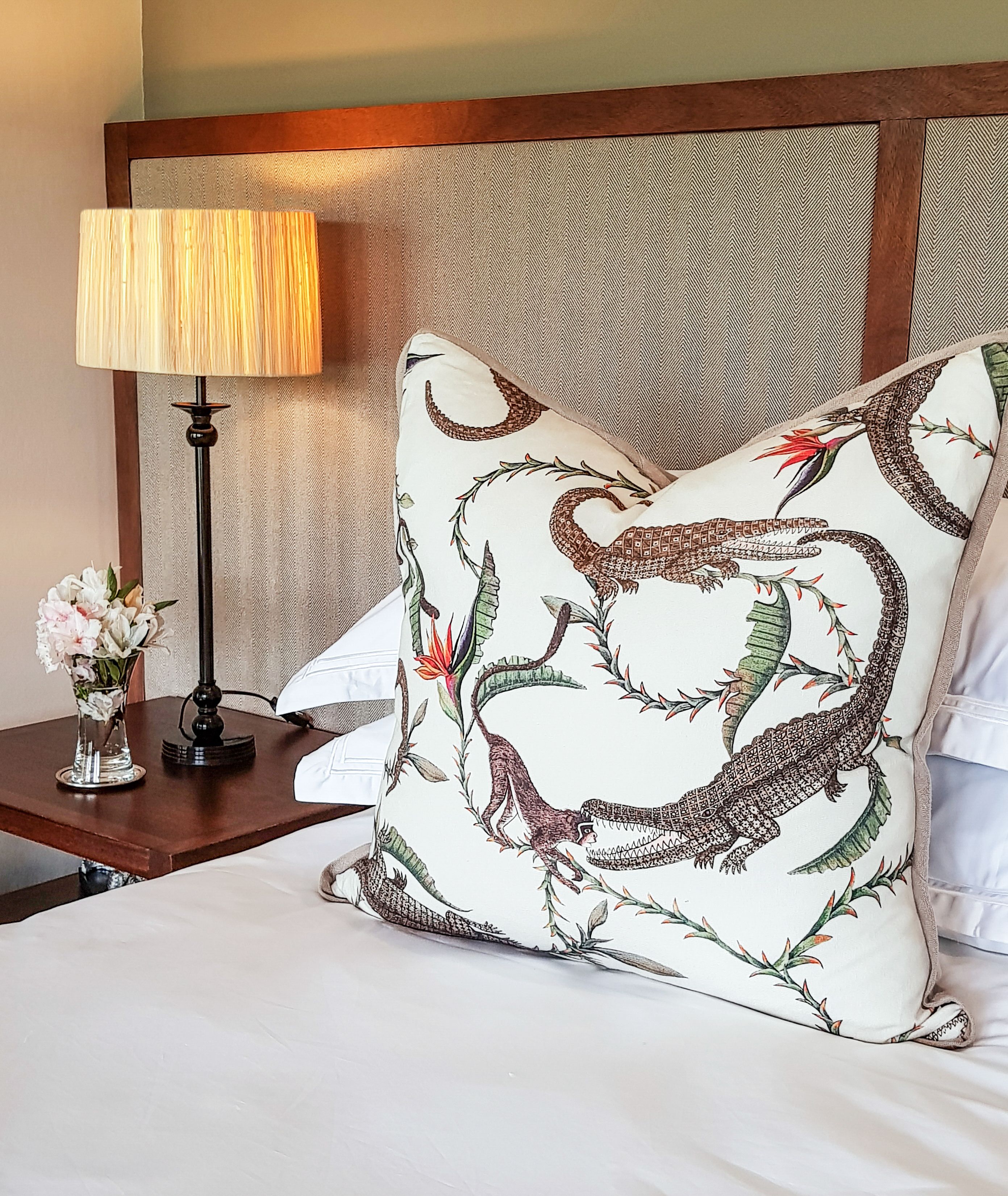 Una cama bien hecha con un almohadón grande de diseño con motivos de reptil, una lámpara de noche y un pequeño jarrón con flores sobre una mesita de noche de madera.
