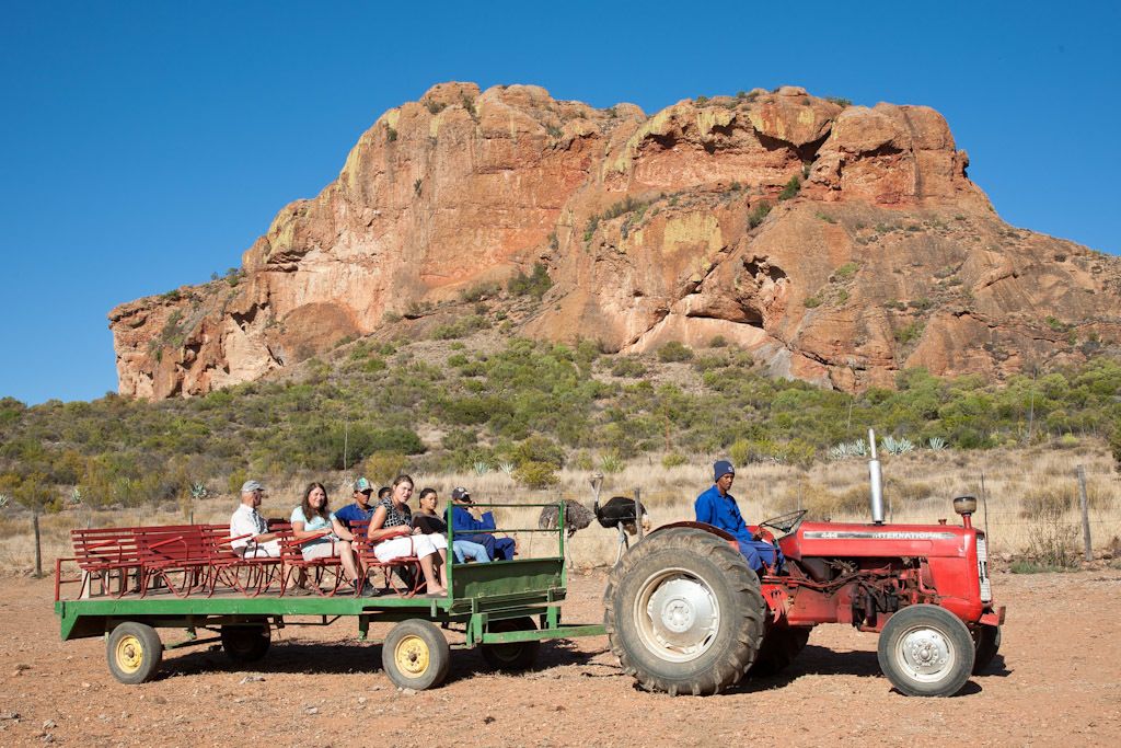Un grupo de personas en un tractor rojo que tira un remolque verde con plataforma, en un paisaje desértico con una gran formación rocosa al fondo.