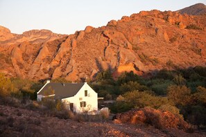 A white house with a sloped roof, surrounded by dry vegetation and rocky hills.