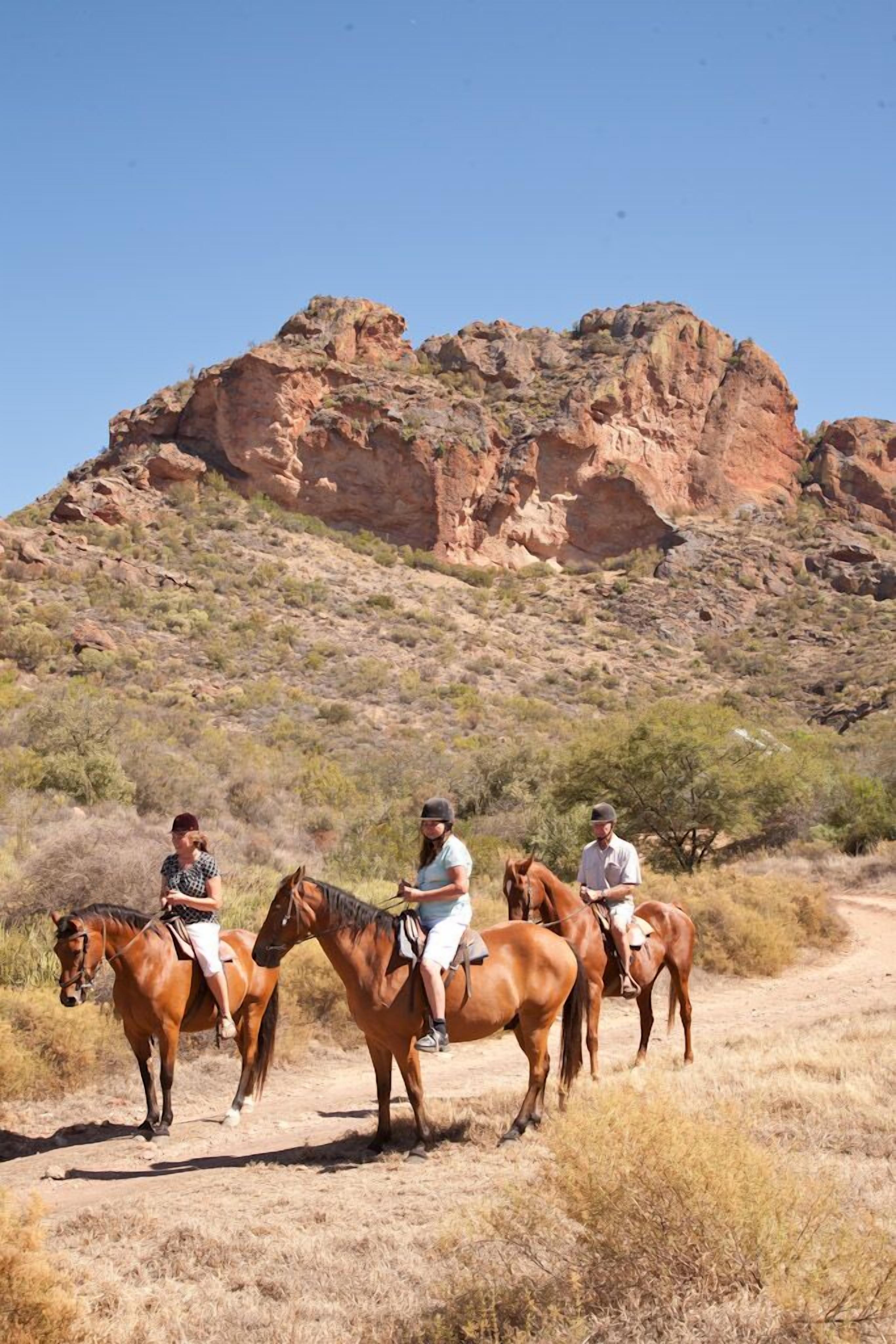 Un grupo de personas montando a caballo por un sendero de tierra con una montaña rocosa al fondo.