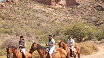 Three people riding horses on a dirt path with a mountainous backdrop.