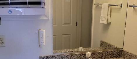 Une salle de bain avec un lavabo doté d’un plan de travail en granit, un miroir, un porte-serviettes, une fenêtre avec des stores et une chaise.