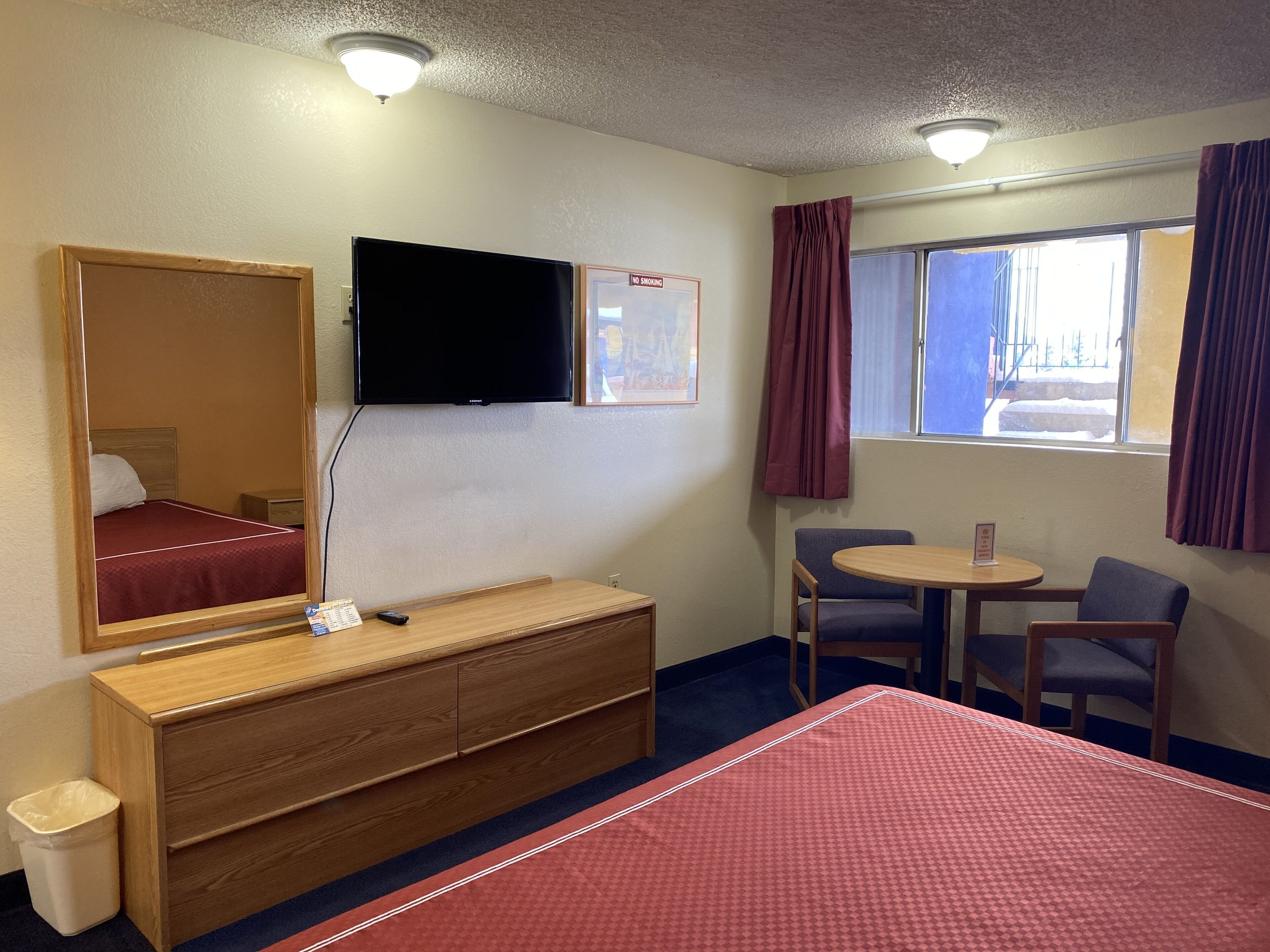 A hotel room with a bed, a wooden dresser, a TV mounted on the wall, a small table with two chairs, and a window with curtains.