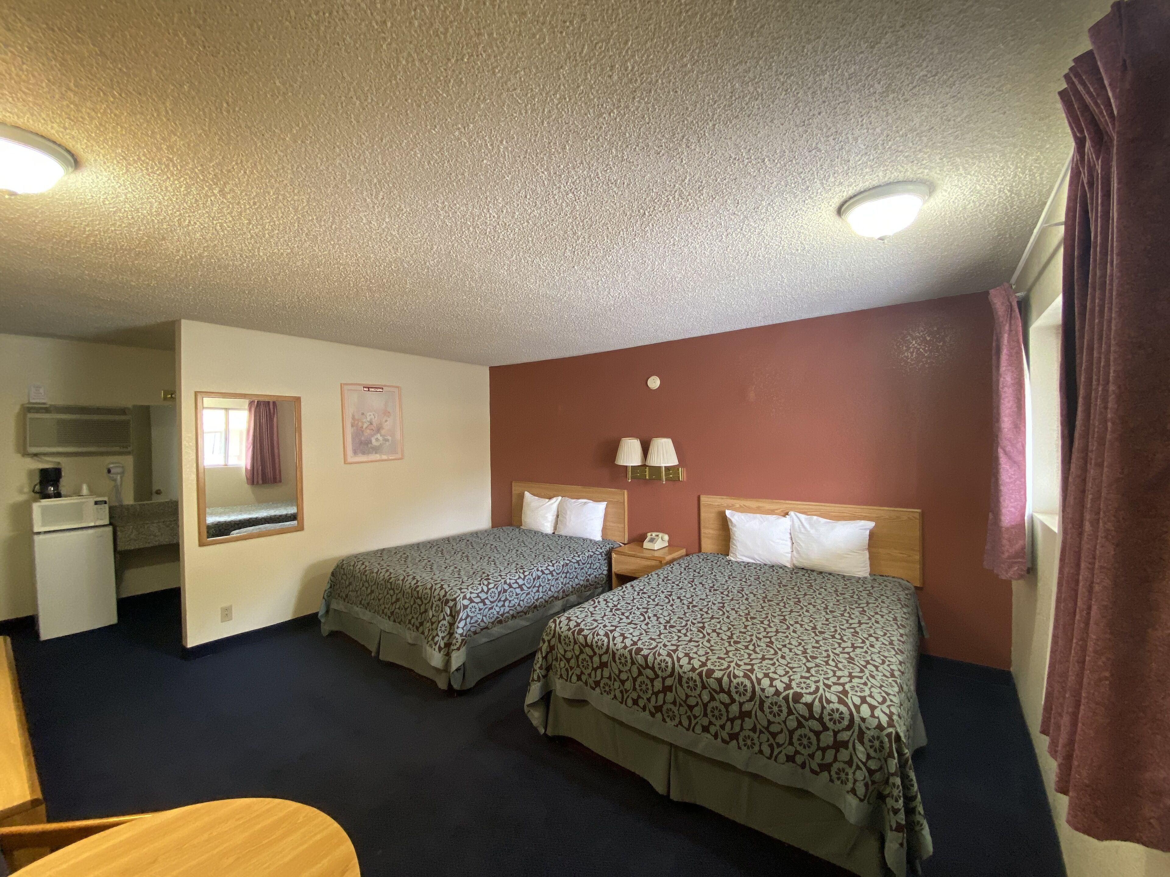 A hotel room with two beds, a desk, a mirror, a wall-mounted TV, and a window with curtains.