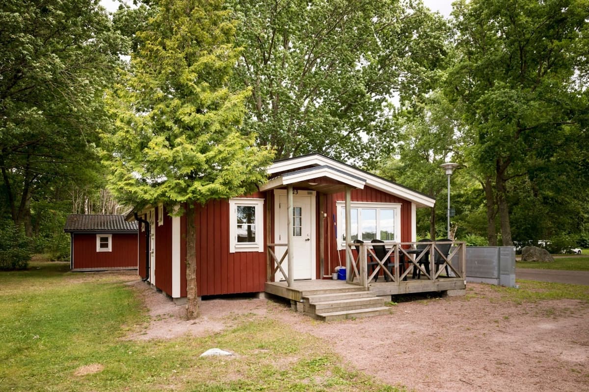 A small, single-story cabin with a porch and a small attached shed, surrounded by trees.