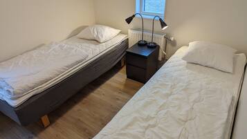 Ein kleines, sauberes Zimmer mit zwei Einzelbetten, einem Nachttisch mit Lampe und einem Fenster mit Tageslicht.
