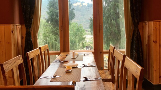 Una mesa de comedor de madera dispuesta con cubiertos y un cuenco de fruta, flanqueada por sillas, con vistas a un paisaje montañoso a través de puertas abiertas.
