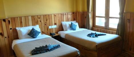 Una habitación con dos camas de madera, cada una con una almohada y una toalla doblada. Hay una ventana con cortinas y una lámpara fijada a la pared.
