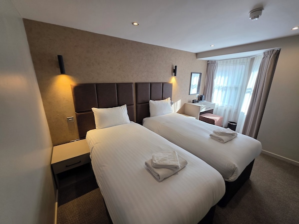 A hotel room with two beds, a nightstand, a desk, and a window with curtains. - Quarterz (Chester)