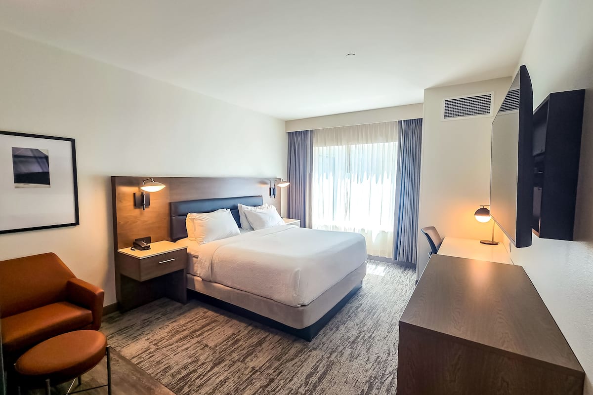 Quarto de hotel com cama, escrivaninha, cadeira, televisão e um quadro pendurado na parede.