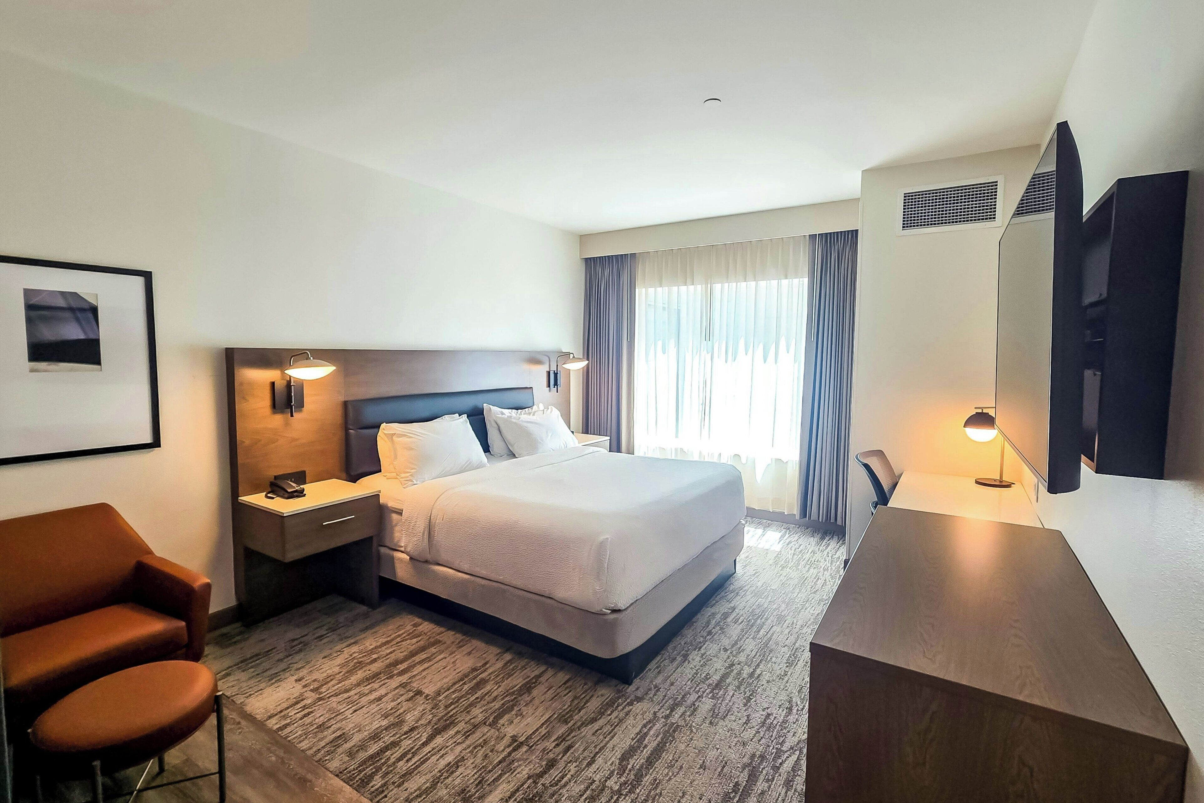 A hotel room with a bed, a desk, a chair, a TV, and a framed picture on the wall.