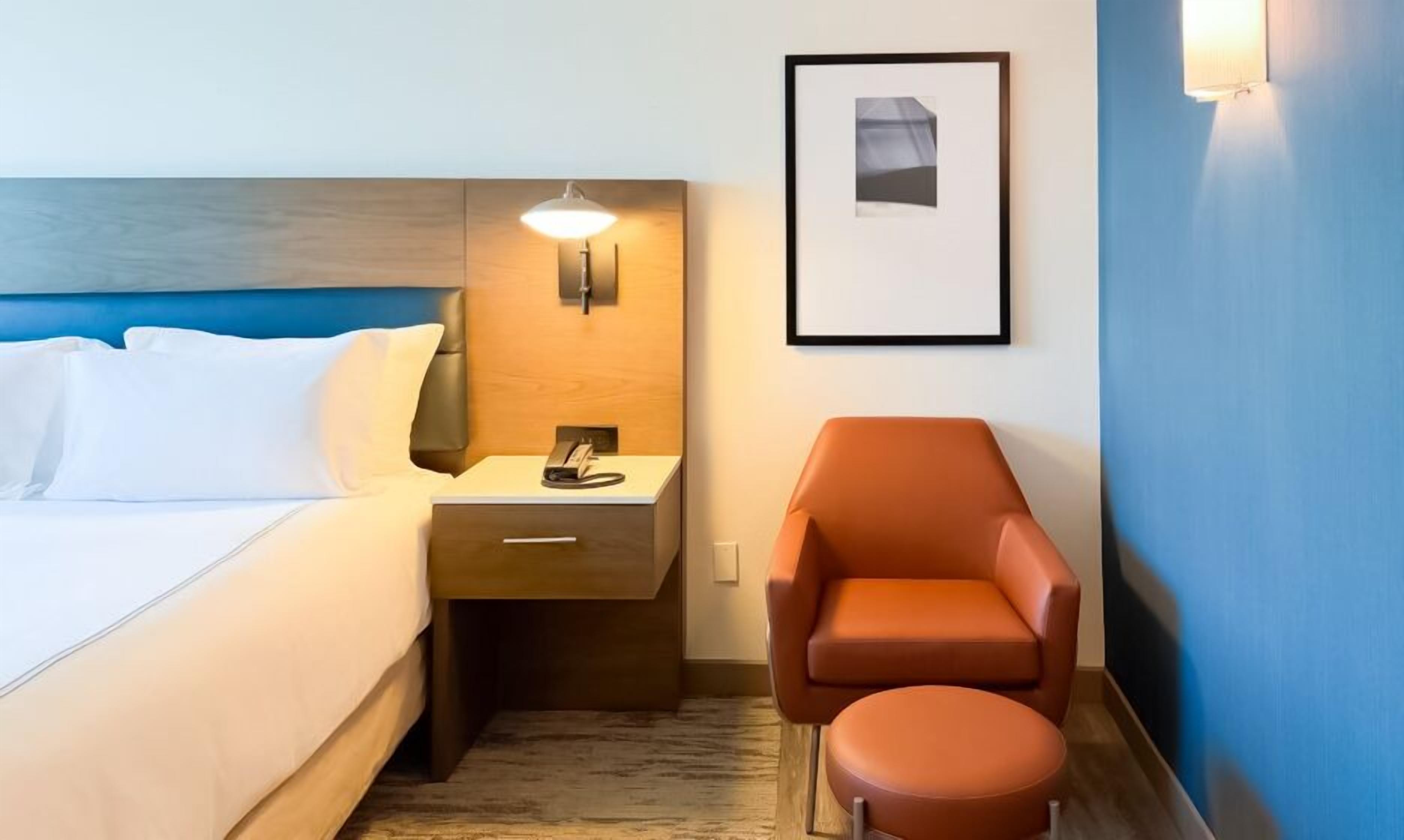 Une chambre d’hôtel avec un lit, une table de chevet, une lampe, une chaise et un tableau encadré au mur.
