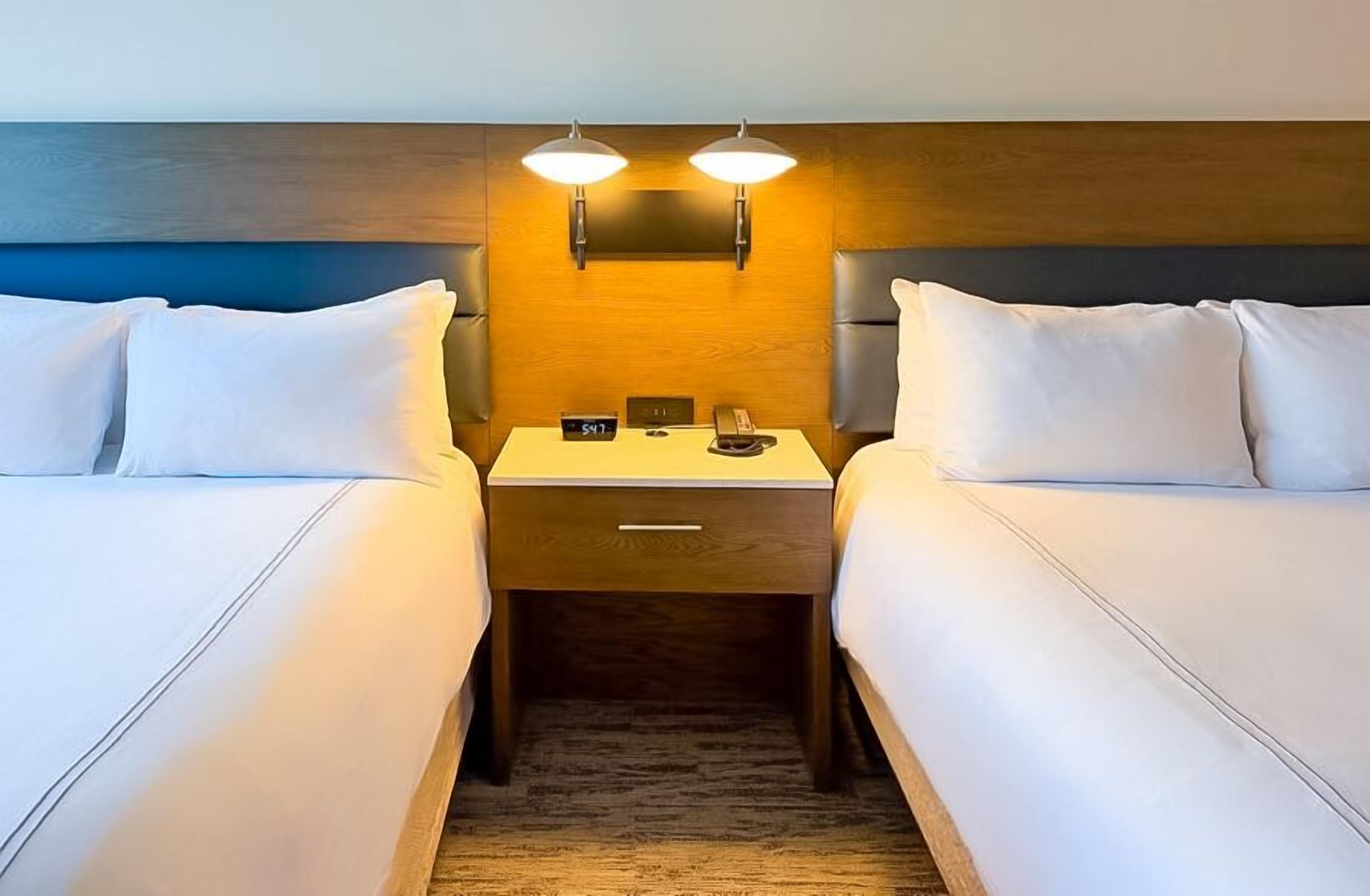 A hotel room with two beds, a nightstand with a clock and a phone, and wall-mounted lights.