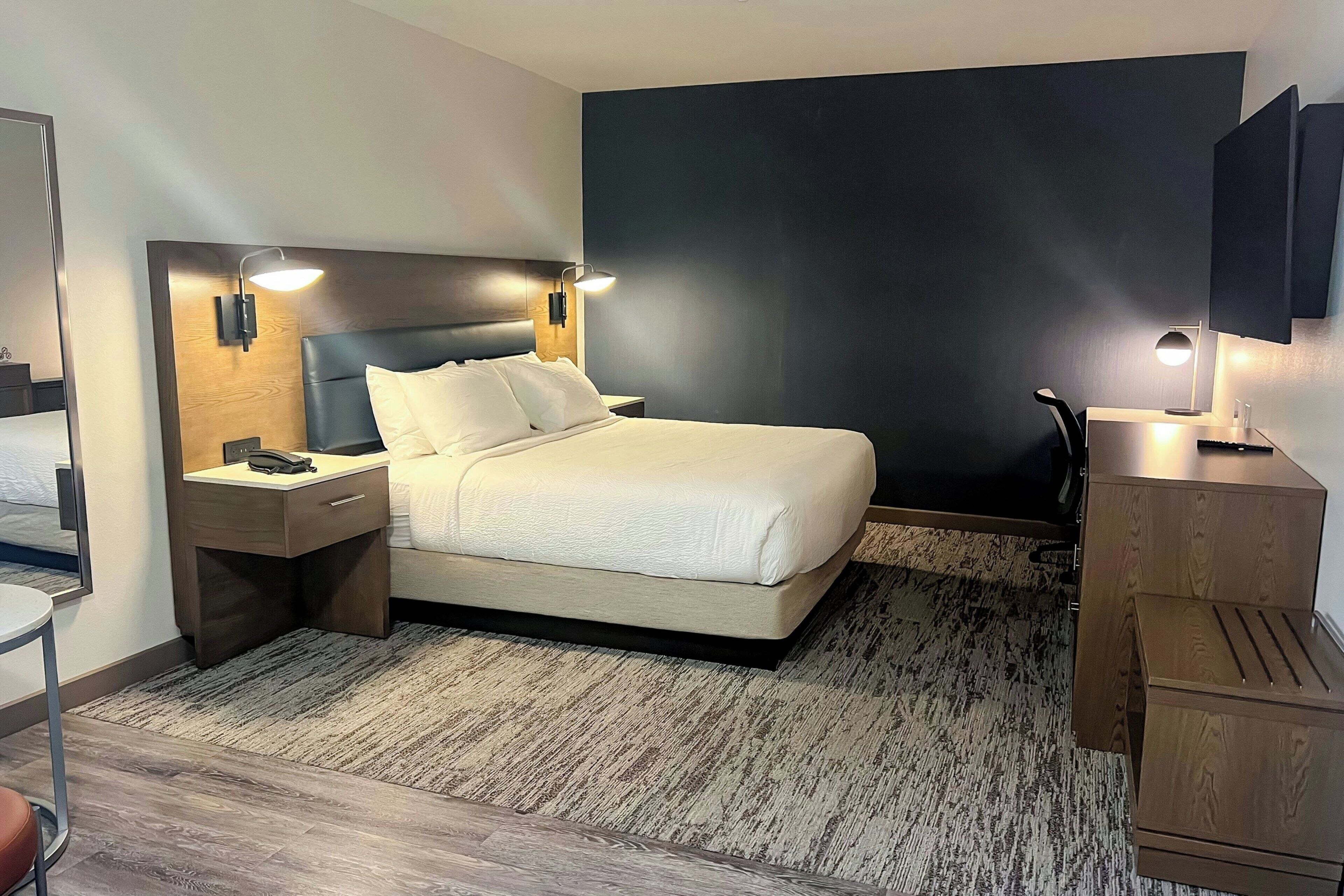 A hotel room with a bed, bedside tables, a desk, a chair, a TV, and a large dark wall.