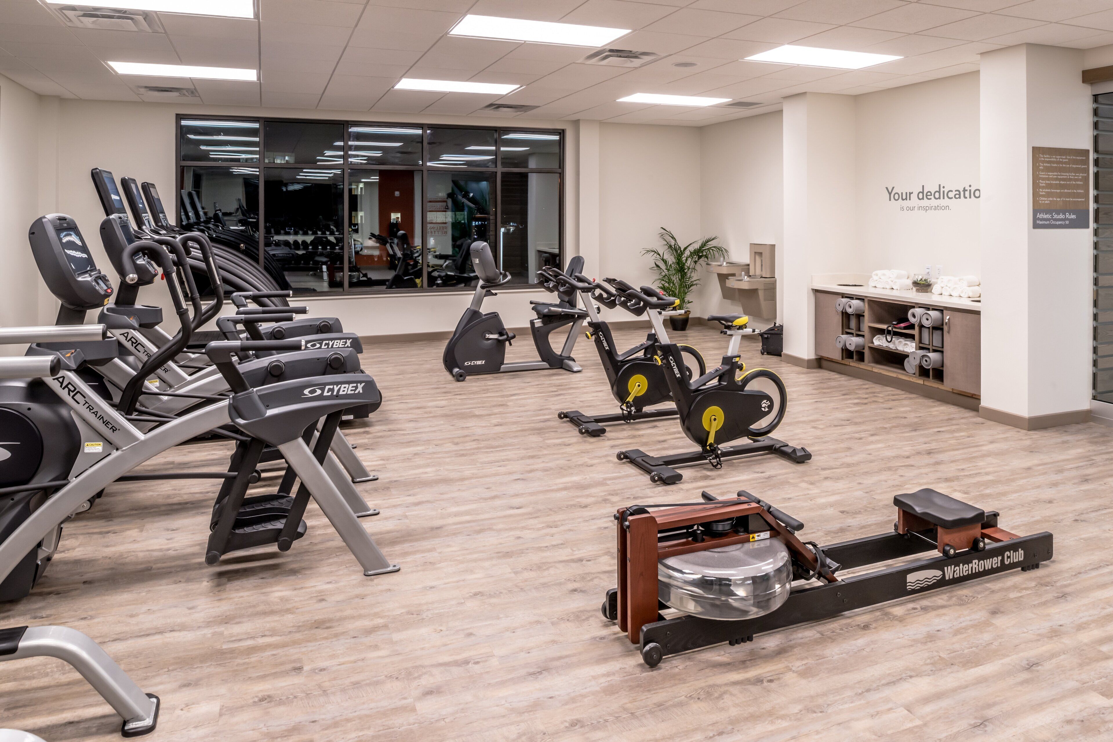 A gym with cardio machines, a rowing machine, and a reception area with a sign.
