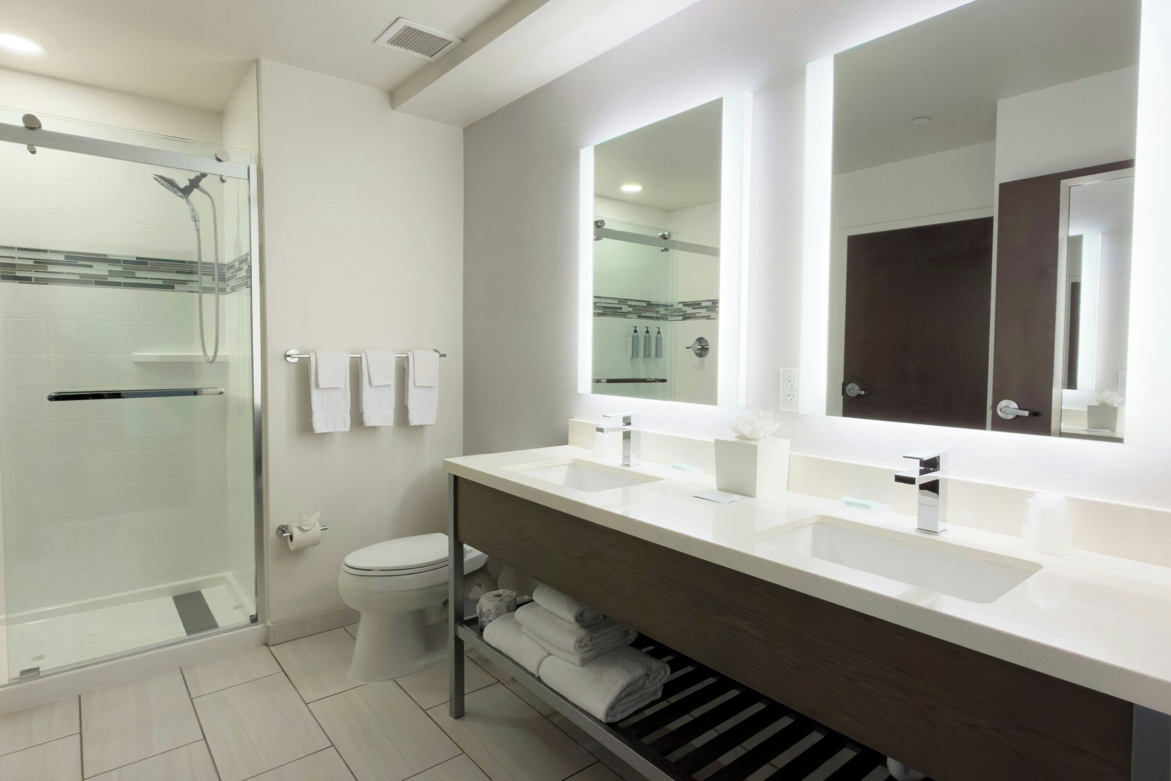 Een moderne badkamer met een douchecabine van glas, een toilet, een wastafel met twee wasbakken en een grote spiegel.