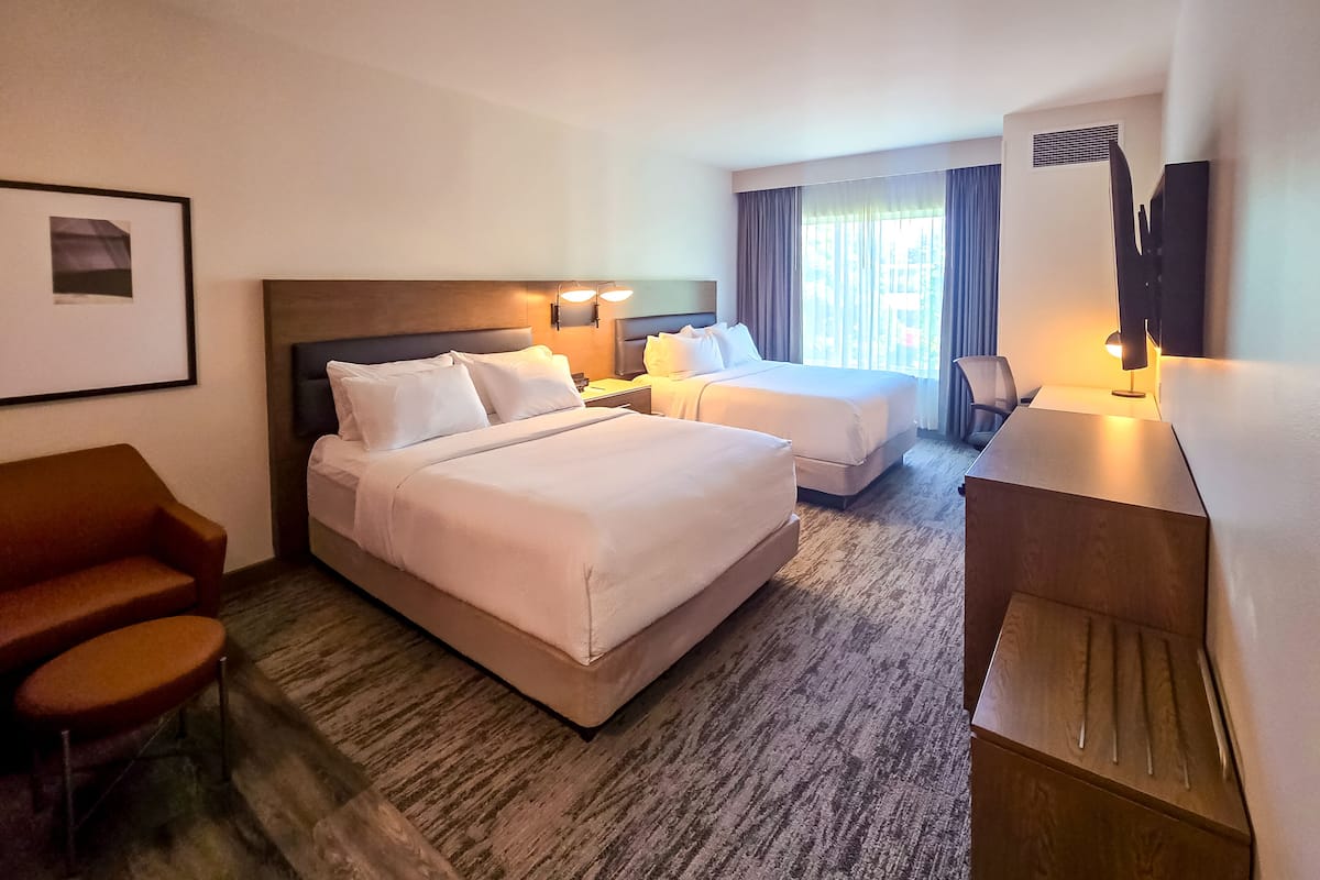 Quarto de hotel com duas camas, uma escrivaninha, uma cadeira, um sofá e uma televisão.