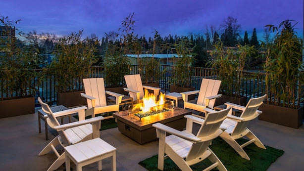 A fire pit surrounded by white Adirondack chairs on an outdoor patio with a view of trees and a building in the background. - Courtyard by Marriott Eugene Downtown (Eugene)